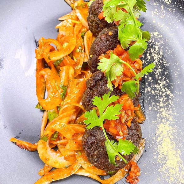 Indian dish with dark meatballs, orange-sauced noodles, and fresh cilantro on a dark plate.