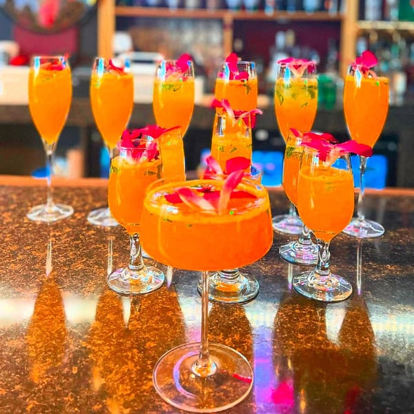 Several orange cocktails garnished with pink petals and green herbs on a dark bar top.