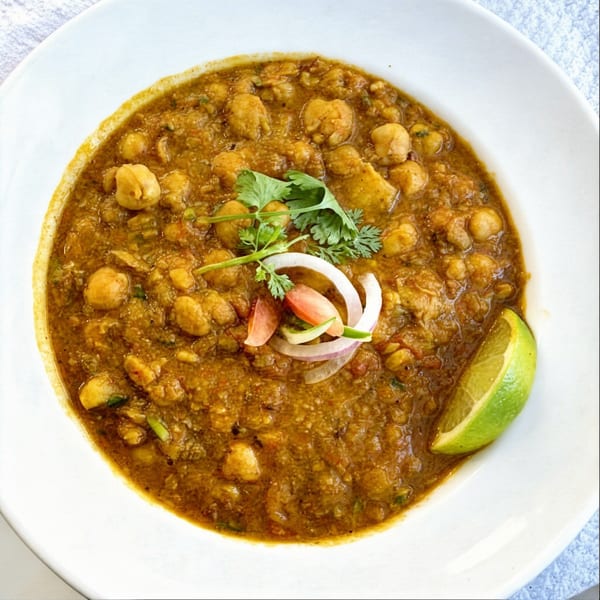 A bowl of chana masala, garnished with cilantro, sliced onions, tomatoes, and a lime wedge.